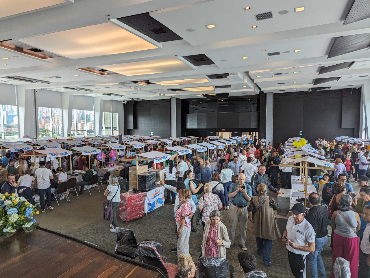 Invoer herverkoop grensoverschrijdende EC is 'n grootskaalse binnenshuise geleentheid met 'n groot aantal mense wat staan en gesels rondom 'n tafel met 'n verskeidenheid items wat in 'n kamer met groot vensters en stadsuitsigte uitgestal word.